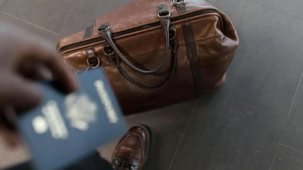 A leather duffel bag on the floor beside a person holding a passport.