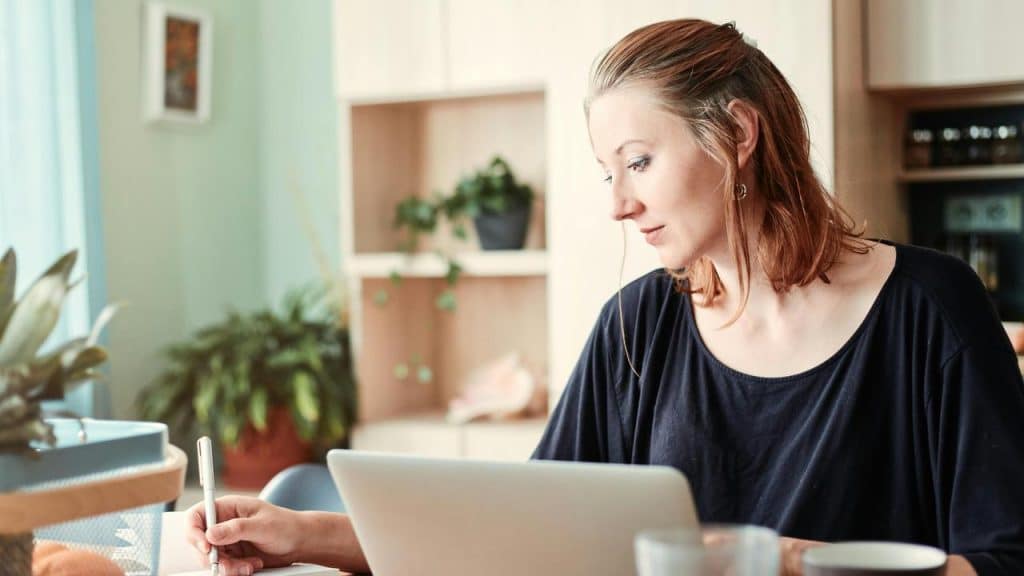 A woman working on a laptop while writing notes at home.