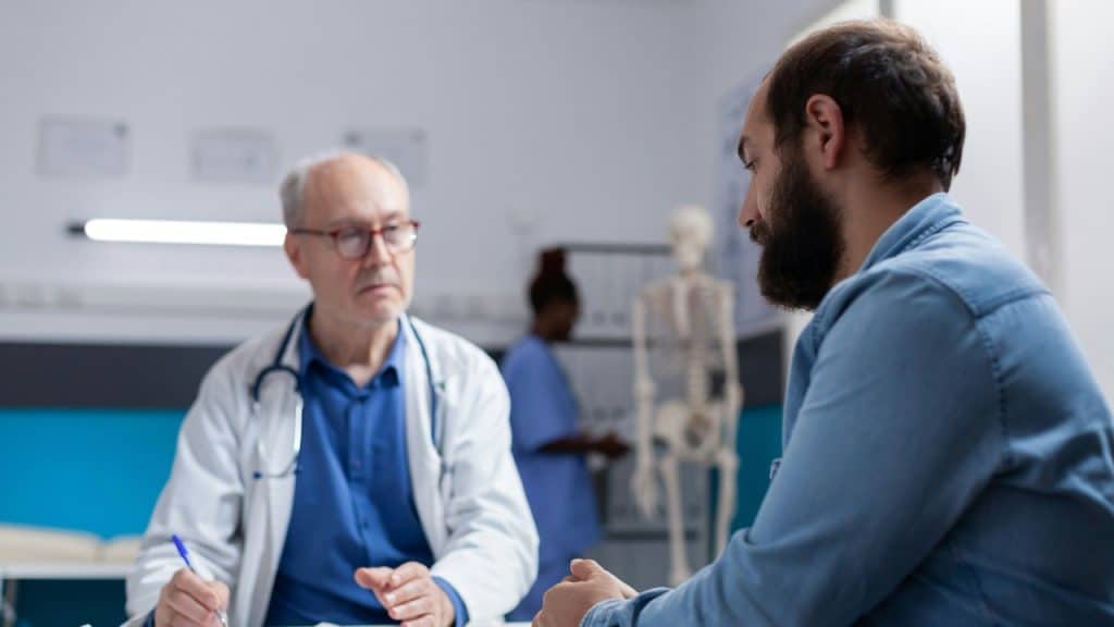 A man with a beard sits across from a doctor in a white coat and glasses.