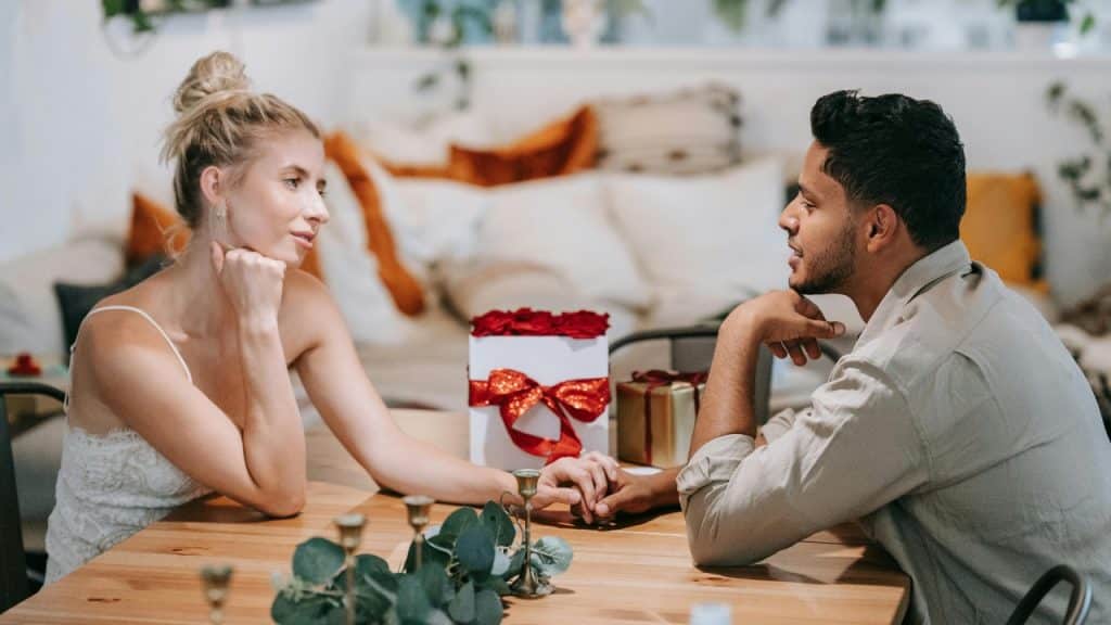 A couple sitting at a wooden table holding hands, with a gift box nearby.