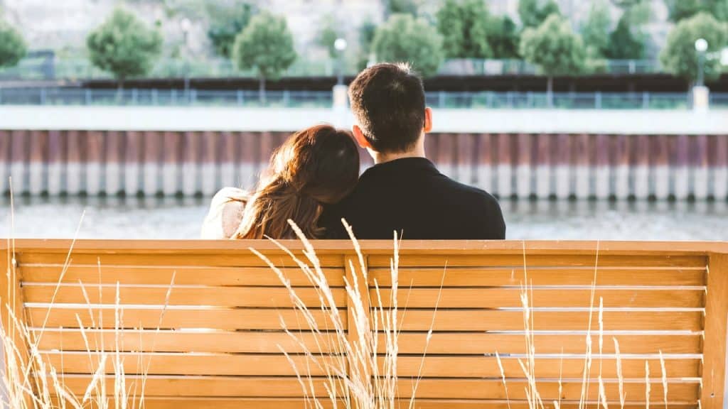 A couple sits on a wooden bench from behind, looking out at a body of water.
