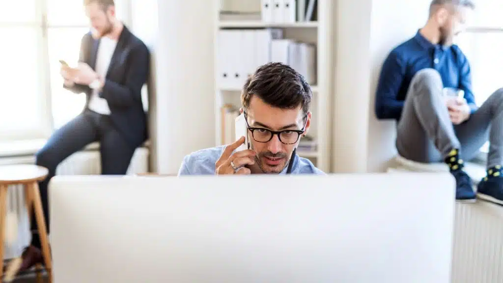 A man talks on a phone behind a computer monitor with two people behind him.