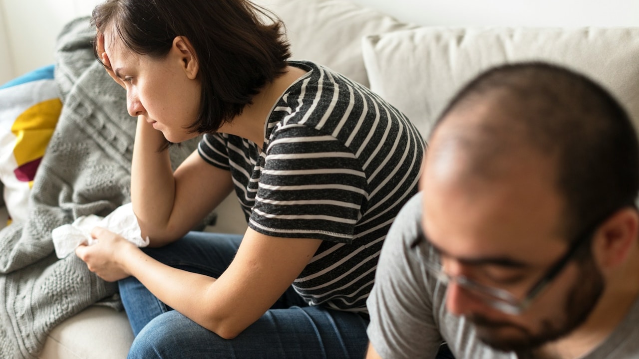 Man and woman sitting on a sofa looking sad, with the woman holding a tissue