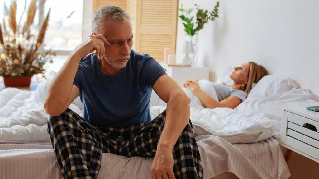 An older man sitting on a bed looking upset