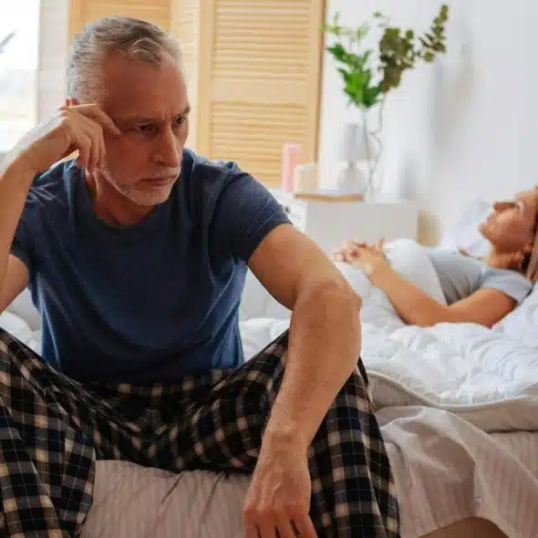 An older man sitting on a bed looking upset