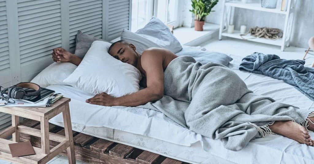 A man sleeping on a bed under a gray blanket in a bright room.