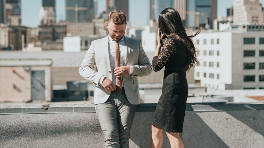 A man in a light suit adjusts his tie while a woman stands nearby.