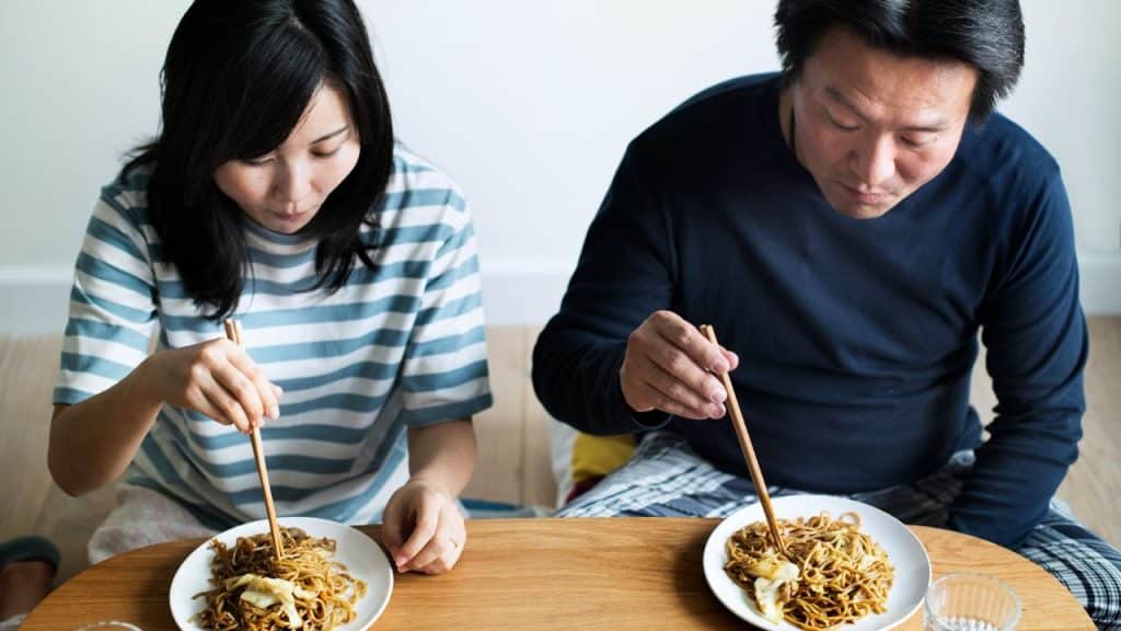A woman and man sit at a wooden table eating noodles with wooden chopsticks.