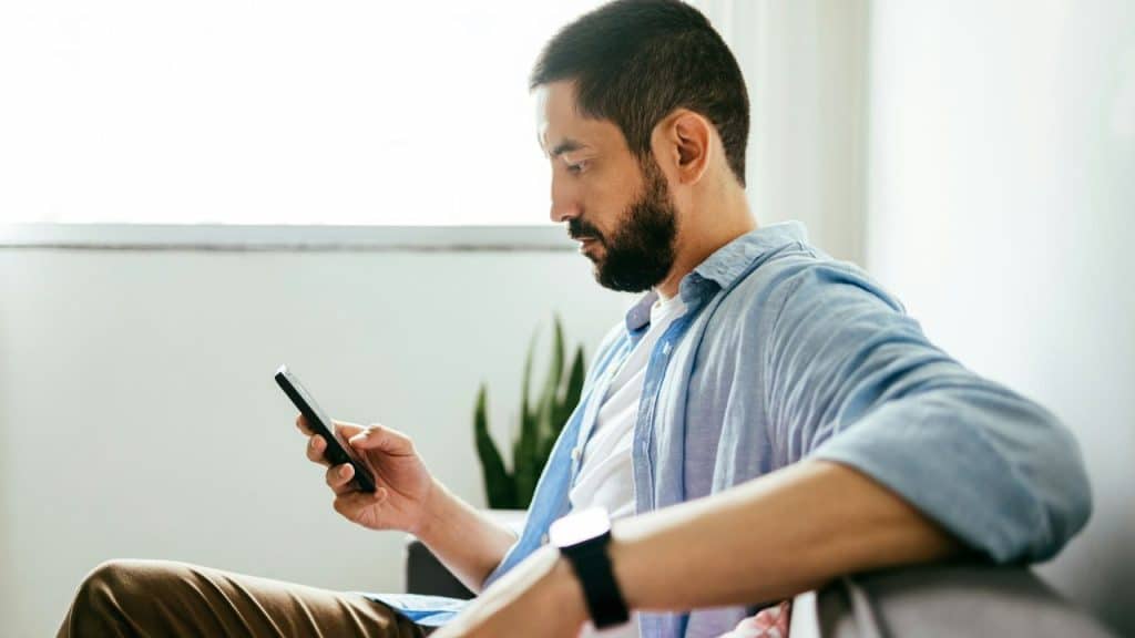 A bearded man in a blue shirt sits on a sofa, looking down at his smartphone.