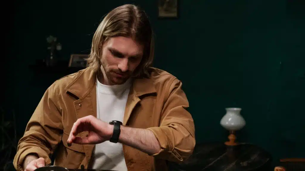 A man with long hair sits in a dark room and looks at his wristwatch.