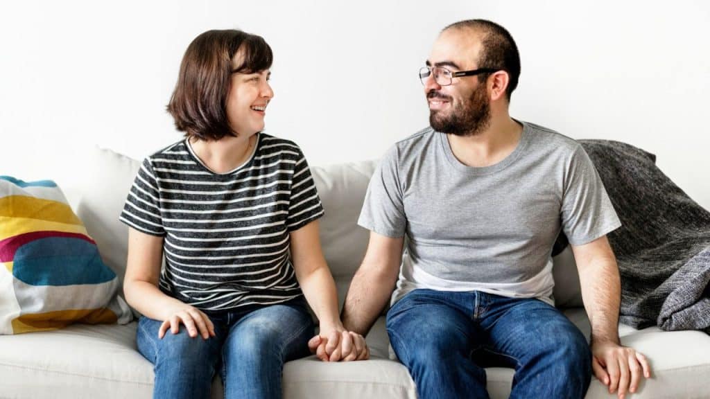 A smiling man and woman sit on a couch together while holding hands and looking.