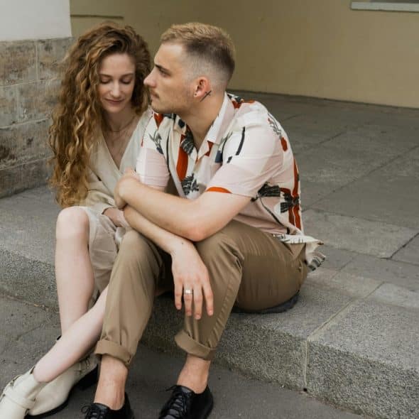 A man and woman sit closely together on concrete steps outside a stone-walled building.