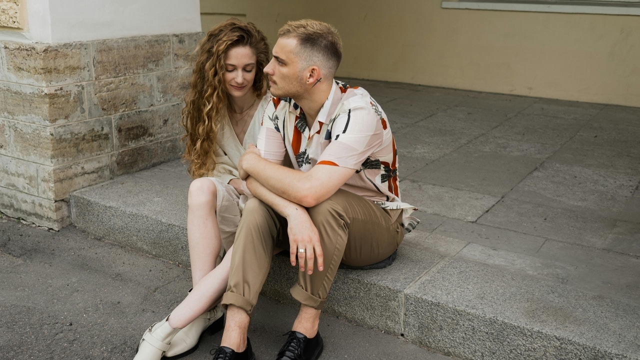A man and woman sit closely together on concrete steps outside a stone-walled building.