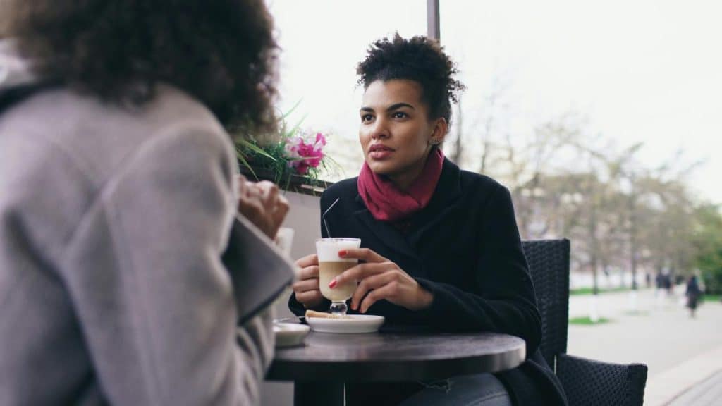 A woman in a scarf talking with someone at an outdoor café table.