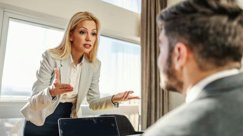 A blonde woman in a blazer gestures with open palms while speaking to a man.
