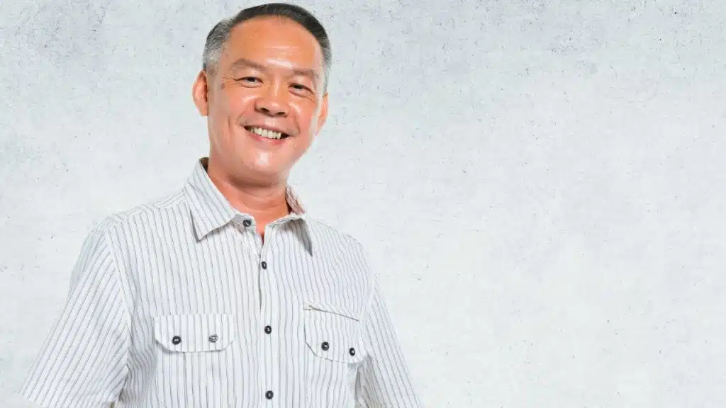 Smiling man with short salt-and-pepper hair wearing a striped button-down shirt against a textured wall.