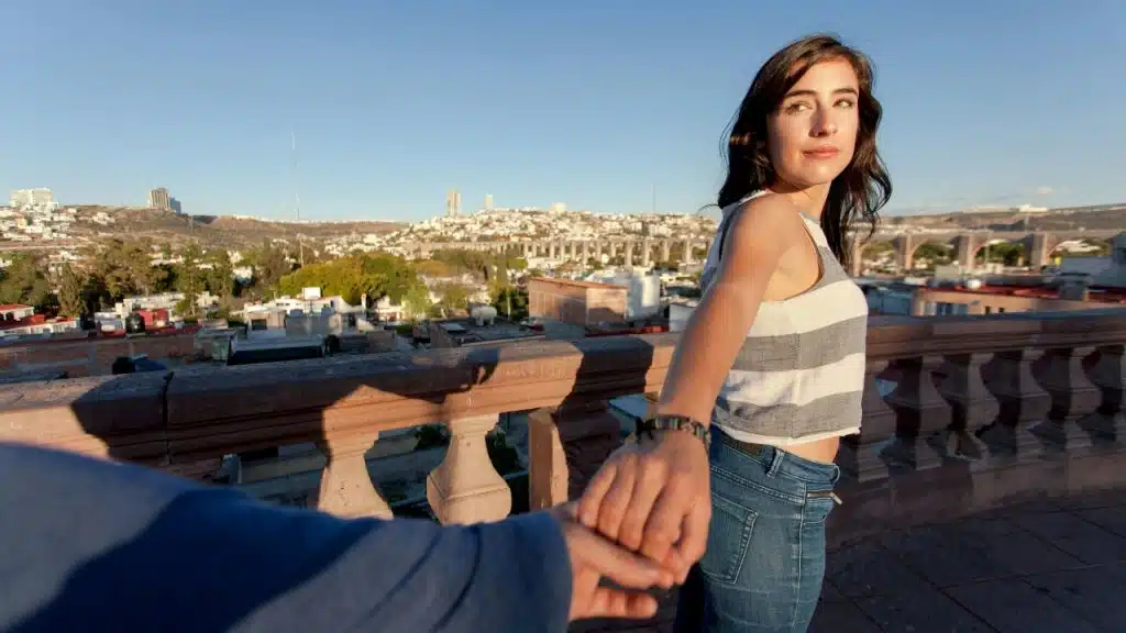 A woman in a striped top holds a hand and looks back over a city balcony.