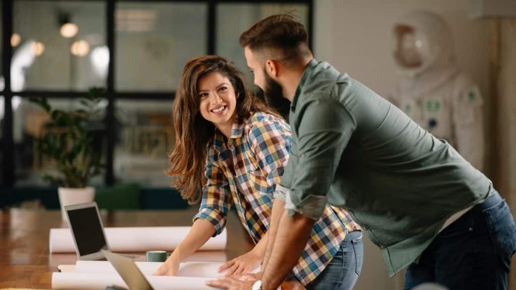 A man and woman look at each other while leaning over large papers on a table.