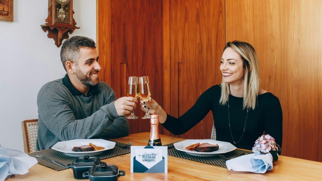 A man and woman sit at a table clinking glasses over a meal with wine.