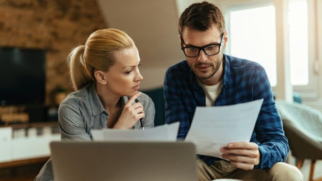 A man and woman sit together indoors while looking at and holding several papers.