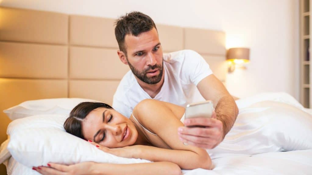 A man with a shocked expression looks at a smartphone while a woman sleeps nearby.