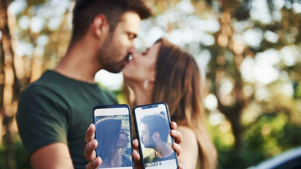 A couple is kissing while having mobile phones in their hands.