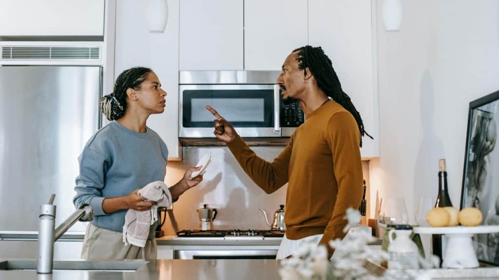 A man in an orange sweater points a finger at a woman in a kitchen.