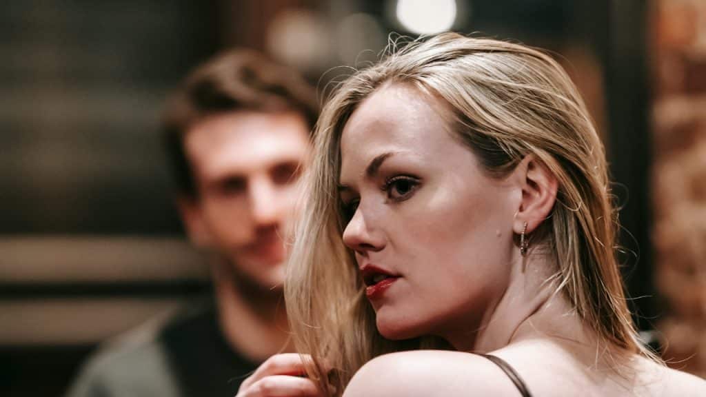 A woman with blonde hair looks off-camera while a man’s face is blurred in the background.