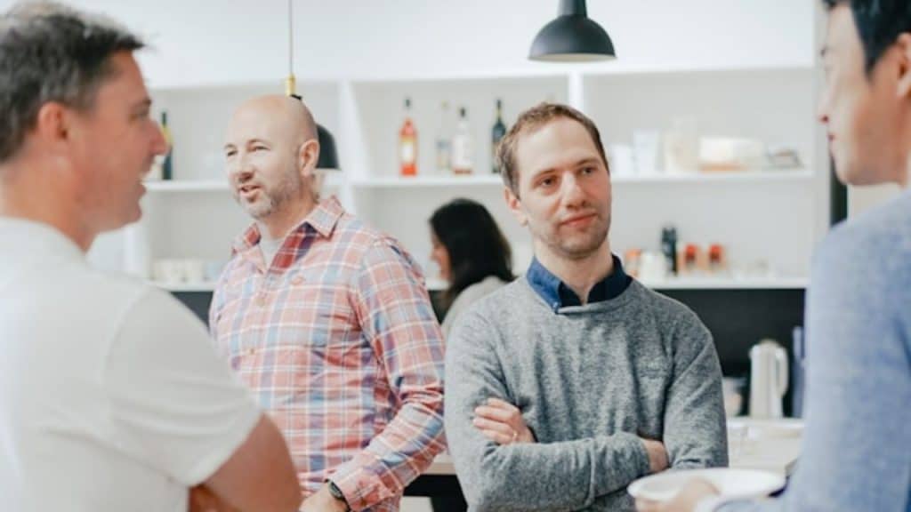 A group of men stand and converse in a brightly lit room with shelves in the background.
