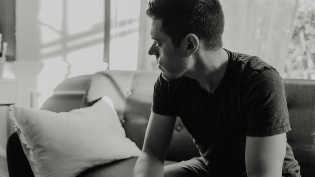 A man sitting on a couch looking to the side, appearing thoughtful in a dim room.