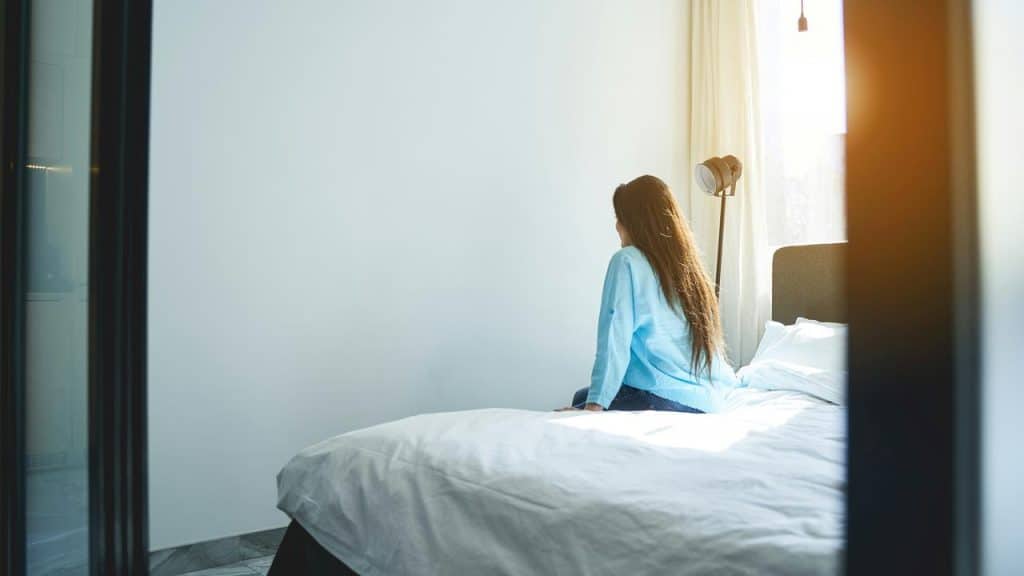 A person sitting on a bed and looking toward a sunlit window.