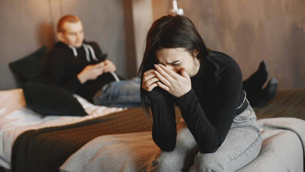 A woman sits on a bed covering her face with her hands while a man watches.