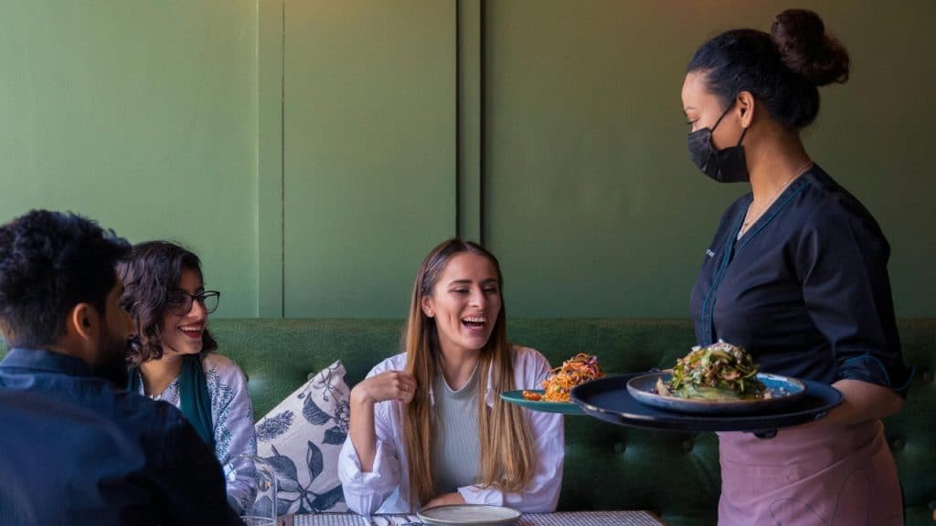 Three people sit at a restaurant booth as a masked server approaches with a tray.