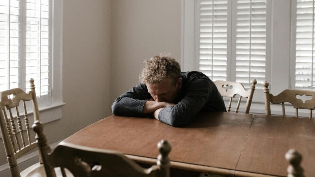 A man with curly hair resting his head on his arms at a wooden table.