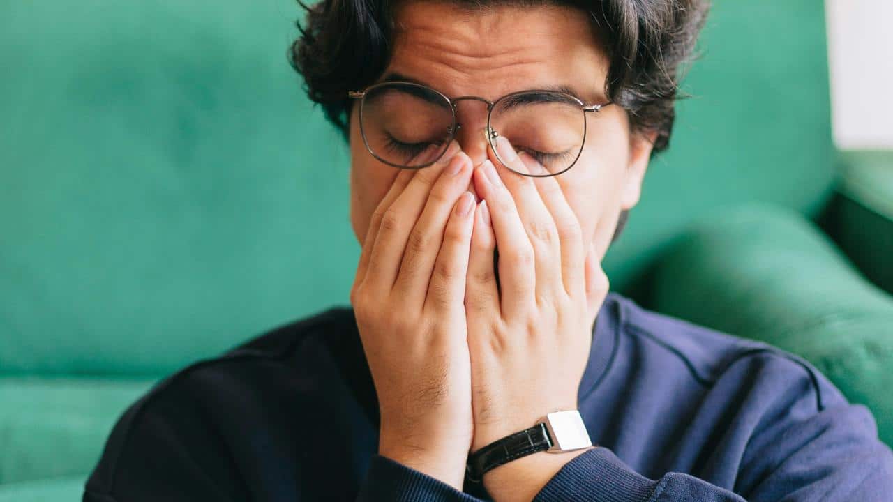 A person with glasses covering their face with their hands