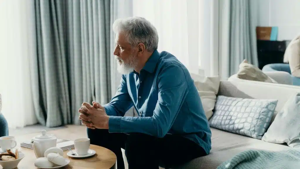 A man with white hair sits on a sofa with his hands clasped together tightly.