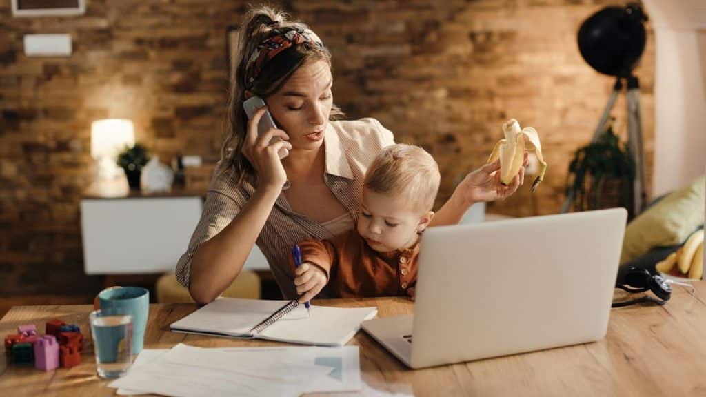 A woman talks on a phone while holding a toddler and a banana at a desk.