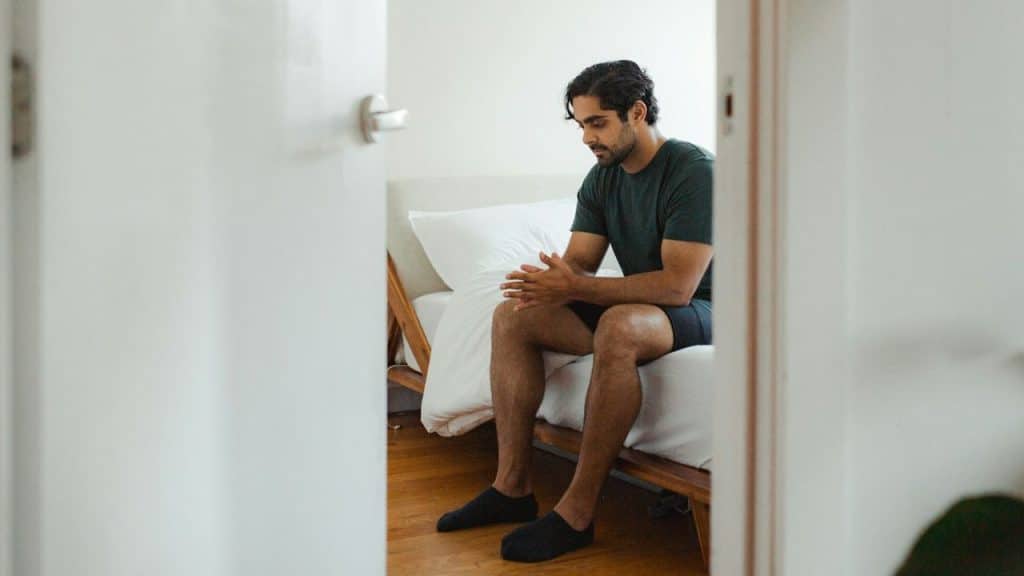 A man sits on the edge of a bed with his hands clasped, viewed through a doorway.