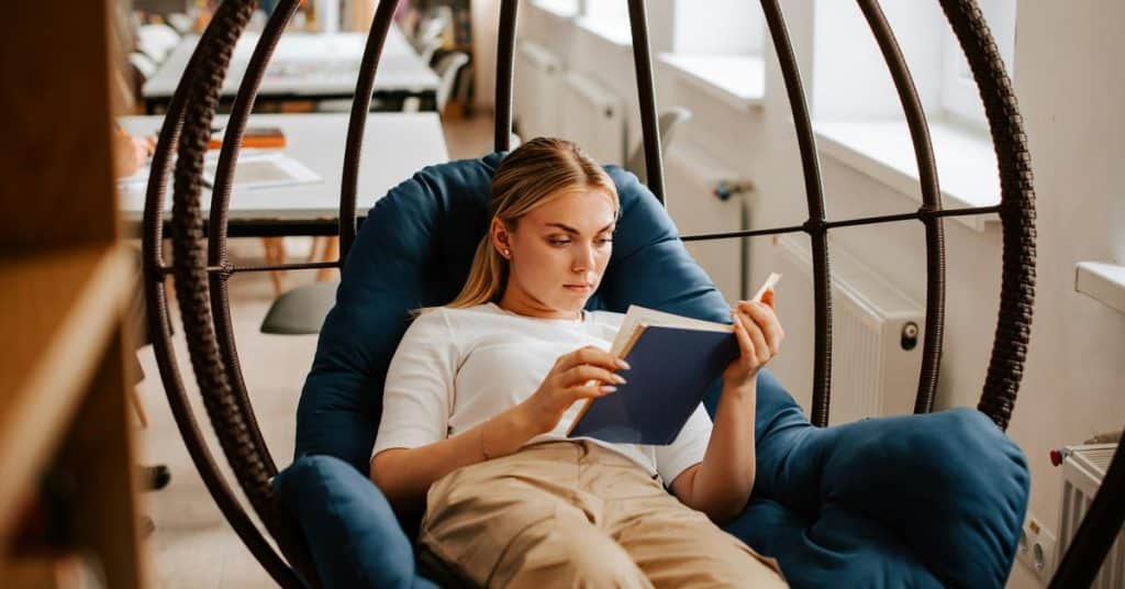 A woman relaxing in a hanging chair while reading a book indoors.