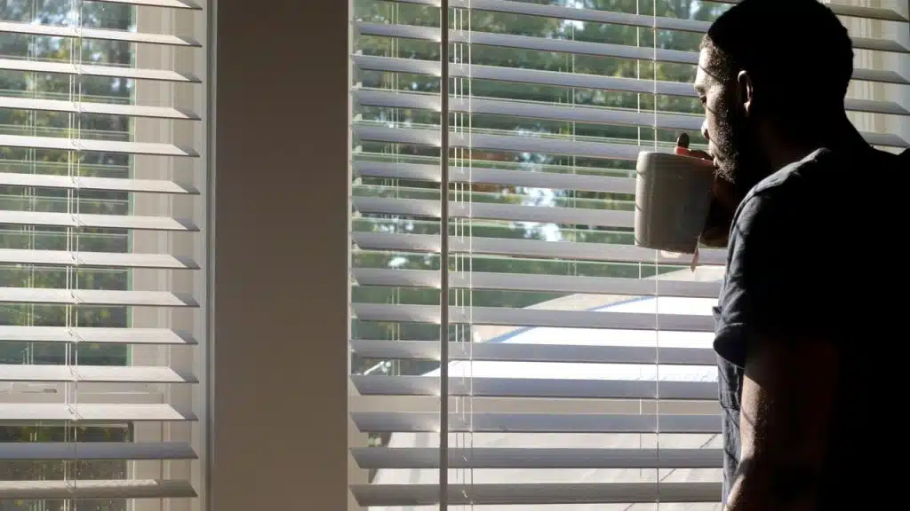 A man drinking from a mug while looking out the window through blinds.