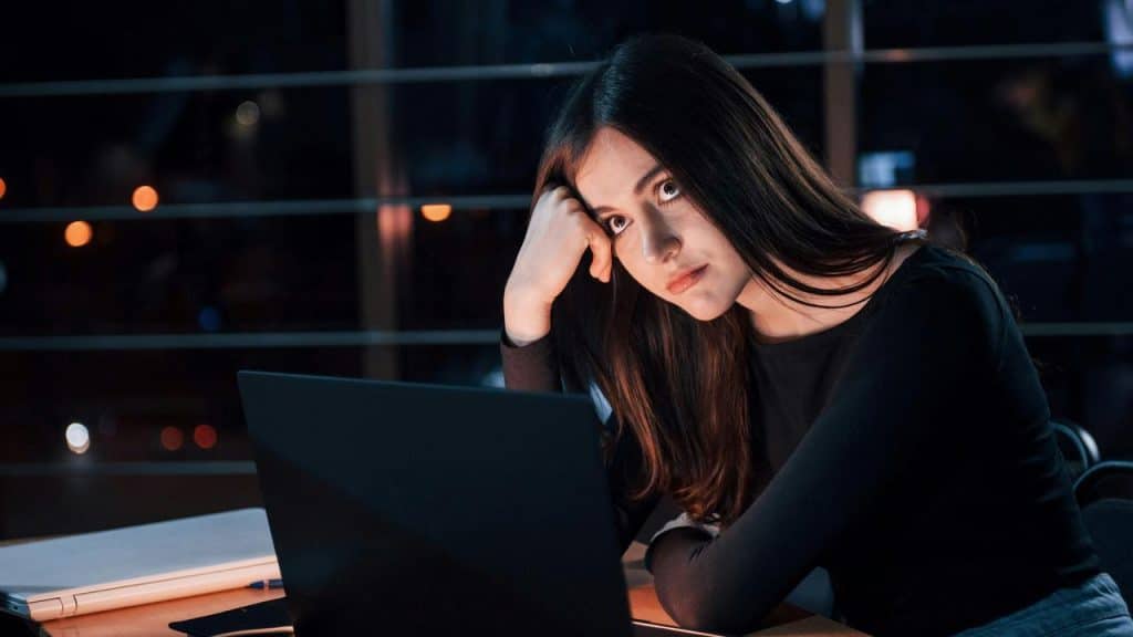 A tired woman resting her head on her hand while working on a laptop at night.