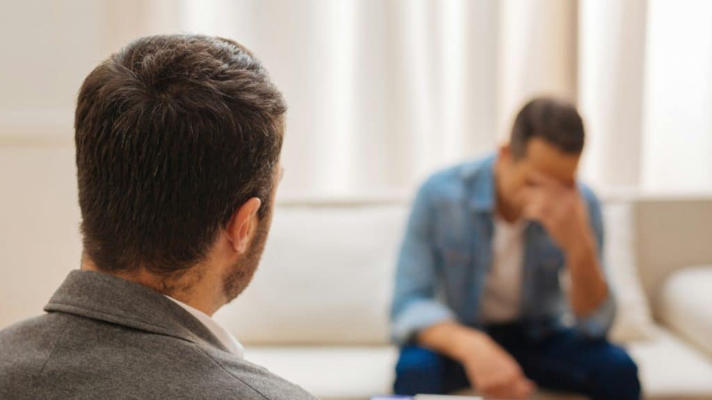 View from behind a man's head looking at another man sitting blurred on a couch.
