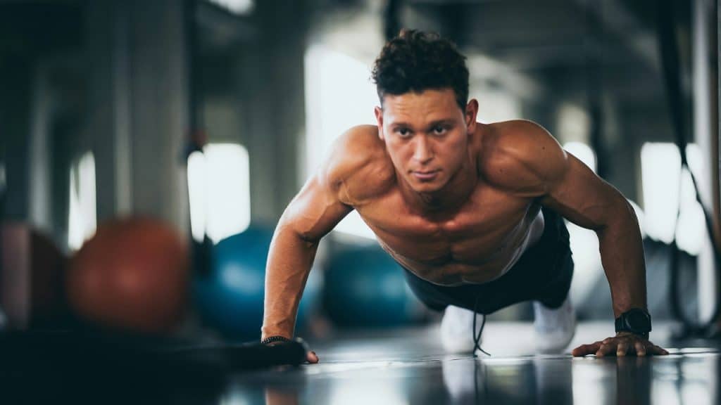 A muscular man performs a pushup on a gym floor while looking directly at the camera.