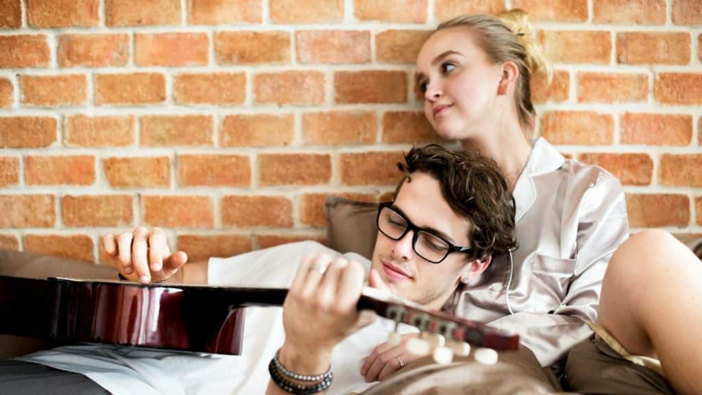 A man lies back playing a guitar while a woman leans against him watching him.