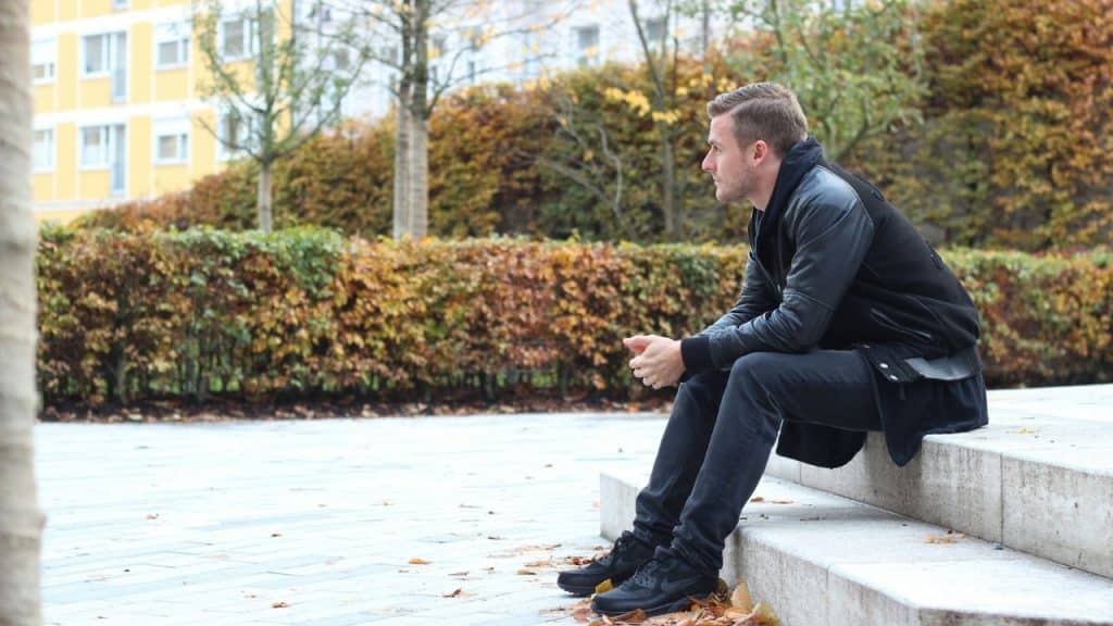 A man in a black jacket sits on concrete steps outdoors near trees with autumn leaves.