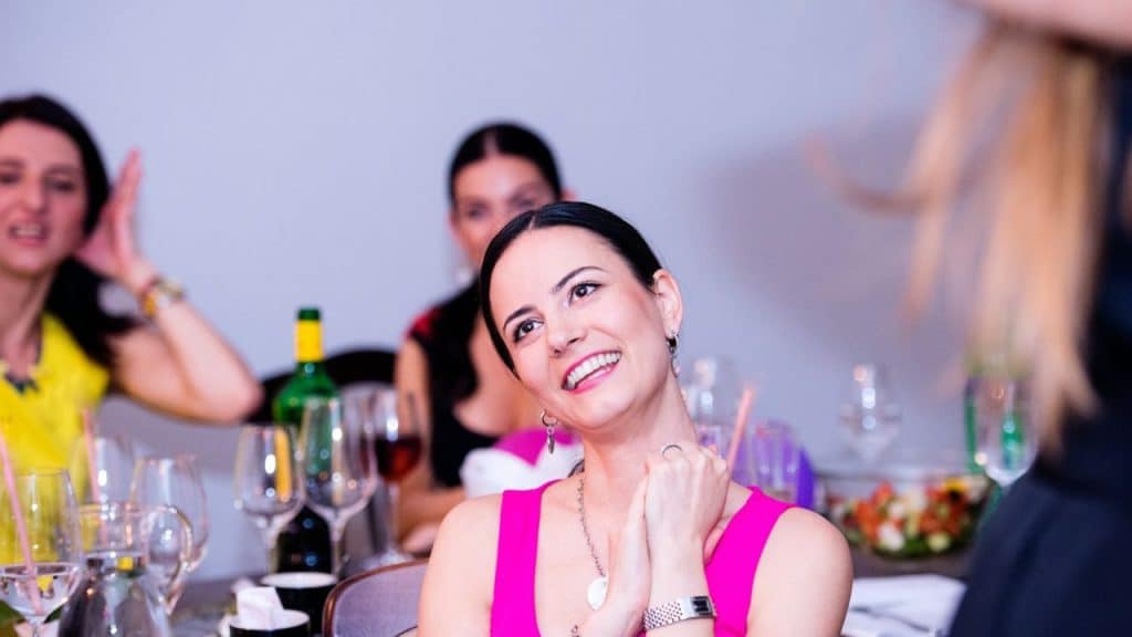 A smiling woman in a pink top is sitting at a table with other people.