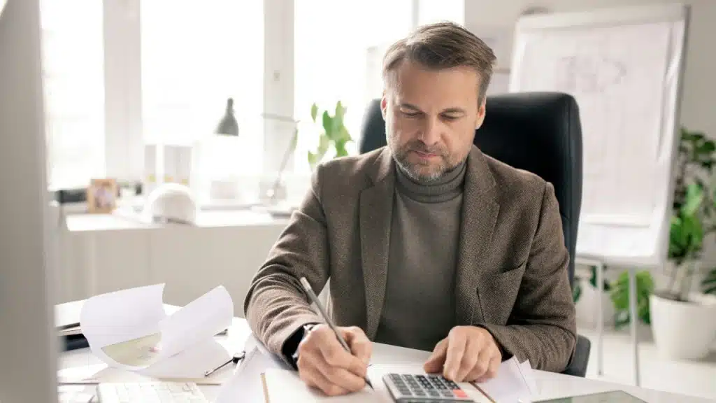 A man in a blazer sits at a desk using a calculator and writing on paper.