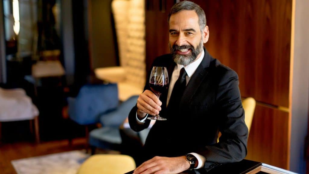 A man in a suit smiles while holding a glass of wine at a bar.