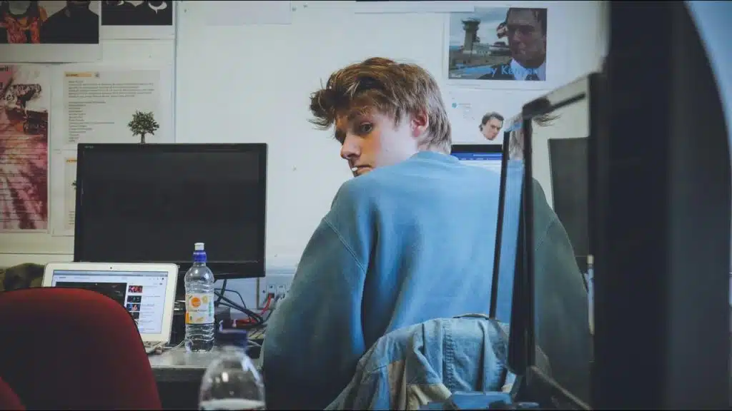 A man turning to look back while sitting at a desk with computer monitors.
