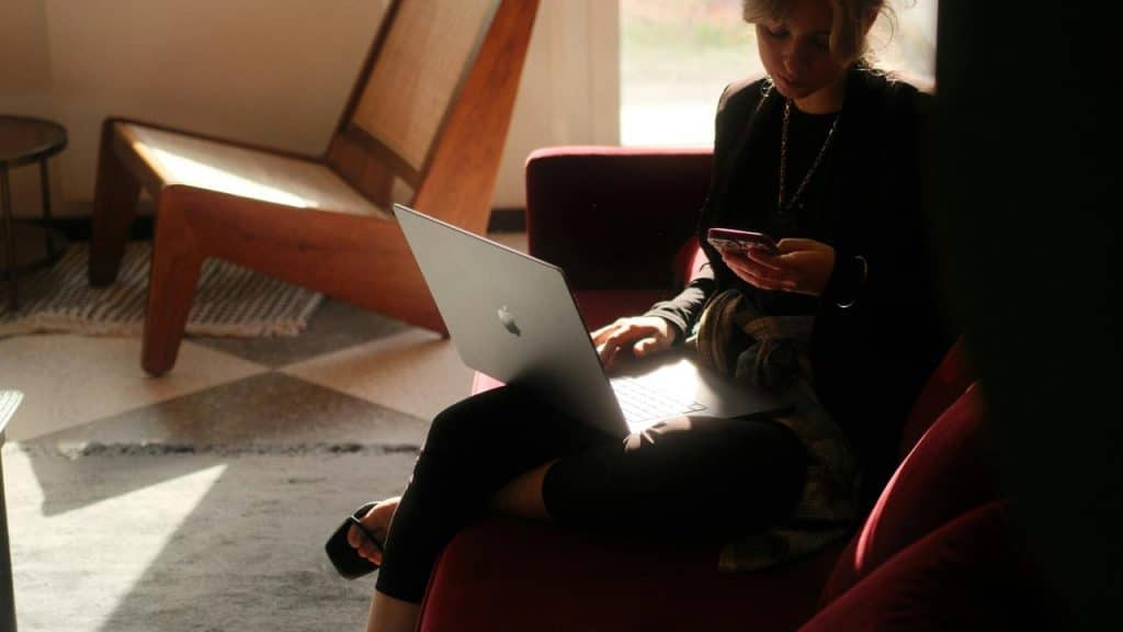 A woman sitting in a chair using a laptop while checking her phone.